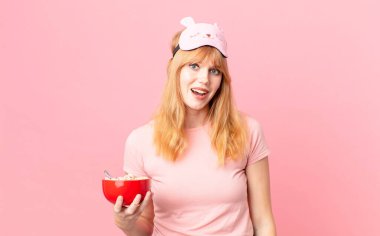 Güzel kızıl saçlı kadın mutlu görünüyor ve hoş bir şekilde şaşırmış pijama giyiyor ve elinde bir kaseyle