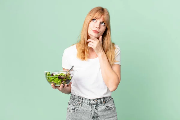 pretty red head woman thinking, feeling doubtful and confused and holding a salad. diet concept