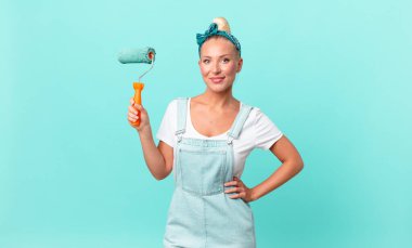 Güzel sarışın kadın mutlu bir şekilde gülümsüyor, eli kalçasında, kendinden emin ve duvar boyuyor.