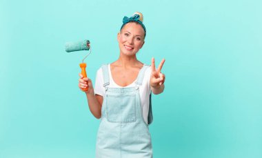 Güzel sarışın kadın gülümsüyor ve mutlu görünüyor, zafer veya barış işareti yapıyor ve bir duvar boyuyor.