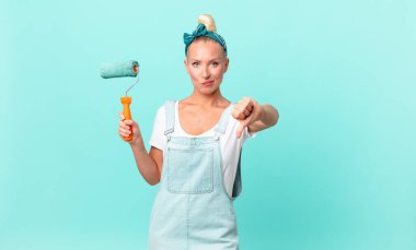 Güzel sarışın kadın, kızgın hissediyor, baş parmaklarını aşağı gösteriyor ve bir duvar boyuyor.