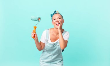 Güzel sarışın kadın mutlu hissediyor, elleri ağızının yanında bağırıyor ve bir duvar boyuyor.