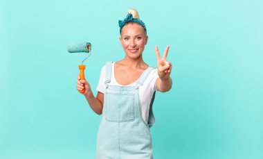 Güzel sarışın kadın gülümsüyor ve arkadaş canlısı görünüyor. İki numarayı gösteriyor ve bir duvar boyuyor.