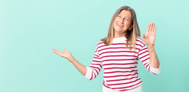 Güzel orta yaşlı kadın mutlu bir şekilde gülümsüyor, el sallıyor, seni selamlıyor ve selamlıyor.