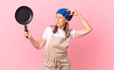 Kafası karışmış, kafası karışmış, başını kaşıyan ve elinde bir tava tutan orta yaşlı bir kadın. Şef konsepti