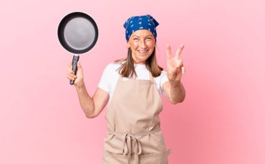 Gülümseyen ve mutlu görünen orta yaşlı bir kadın zafer ya da barış işareti yapıyor ve elinde bir tava tutuyor. Şef konsepti