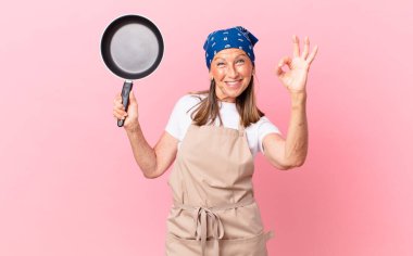 Oldukça orta yaşlı bir kadın mutlu hissediyor, onayını veriyor, iyi bir jest yapıyor ve elinde bir tava tutuyor. Şef konsepti