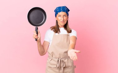 Sevimli orta yaşlı bir kadın mutlu bir şekilde gülümsüyor ve bir konsept sunuyor ve bir tava tutuyor. Şef konsepti