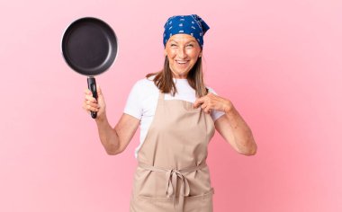 Oldukça orta yaşlı bir kadın kendini mutlu hissediyor ve heyecanlı ve elinde bir tavayla kendini işaret ediyor. Şef konsepti