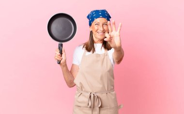 Güzel orta yaşlı bir kadın gülümsüyor ve arkadaş canlısı görünüyor, üç numarayı gösteriyor ve elinde bir tava tutuyor. Şef konsepti