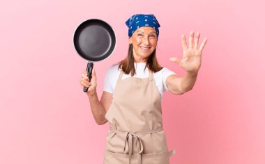 Güzel orta yaşlı bir kadın gülümsüyor ve arkadaş canlısı görünüyor. Beş numarayı gösteriyor ve elinde bir tava tutuyor. Şef konsepti