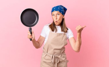 Oldukça orta yaşlı bir kadın inançsızlığa şaşırmış görünüyor ve elinde bir tava tutuyor. Şef konsepti