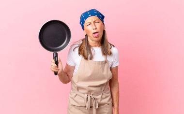 Oldukça orta yaşlı bir kadın, tiksinmiş ve rahatsız olmuş hissediyor ve dilini dışarı çıkarıyor ve elinde bir tava tutuyor. Şef konsepti