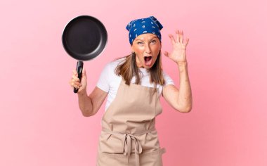 Orta yaşlı güzel bir kadın ellerini havaya kaldırıp tava tutarak çığlık atıyor. Şef konsepti