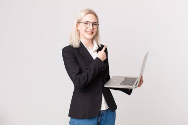 Sarışın, güzel bir kadın neşeli bir şekilde gülümsüyor, mutlu hissediyor ve yan tarafı işaret ediyor ve elinde bir dizüstü bilgisayar tutuyor.