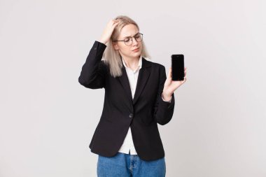 Sarışın, güzel bir kadın kafası karışmış ve kafası karışmış, kaşınıyor ve elinde bir cep telefonu tutuyor.