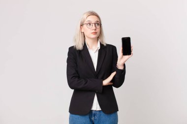 Sarışın, güzel bir kadın omuz silkiyor, kafası karışık ve kararsız ve elinde bir cep telefonu tutuyor.