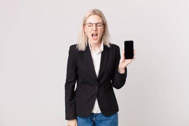 Sarışın, güzel bir kadın agresif bir şekilde bağırıyor, çok kızgın görünüyor ve elinde cep telefonu var.