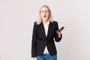 Sarışın, güzel bir kadın agresif bir şekilde bağırıyor, çok kızgın görünüyor ve elinde cep telefonu var.