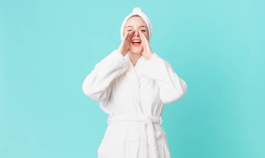 Sarışın, güzel bir kadın mutlu hissediyor, elleri ağızının yanında ve bornoz giymiş olarak bağırıyor.