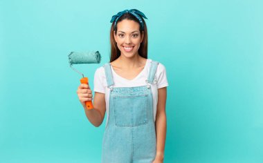 young hispanic woman smiling happily with a hand on hip and confident and painting a wall