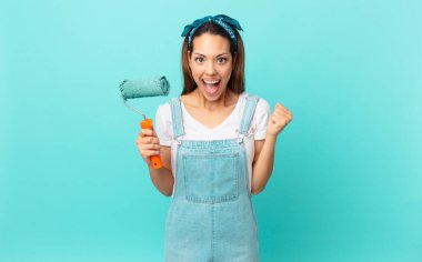 young hispanic woman feeling shocked,laughing and celebrating success and painting a wall