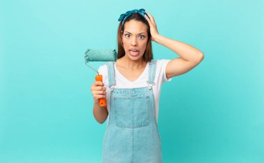 young hispanic woman feeling stressed, anxious or scared, with hands on head and painting a wall
