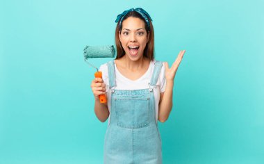 young hispanic woman feeling happy and astonished at something unbelievable and painting a wall