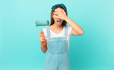 young hispanic woman looking shocked, scared or terrified, covering face with hand and painting a wall