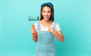 young hispanic woman feeling happy and pointing to self with an excited and painting a wall