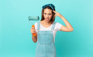 young hispanic woman feeling confused and puzzled, showing you are insane and painting a wall
