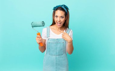 young hispanic woman looking excited and surprised pointing to the side and painting a wall