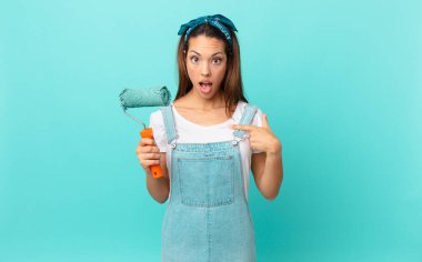 young hispanic woman looking shocked and surprised with mouth wide open, pointing to self and painting a wall