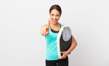 young hispanic woman smiling proudly and confidently making number one and holding a scale
