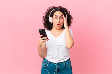 Güzel Arap kadın mutlu görünüyor, şaşırmış ve şaşırmış görünüyor kulaklıklar ve akıllı bir telefon.