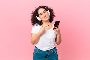 Güzel bir Arap kadını neşeli bir şekilde gülümsüyor, mutlu hissediyor ve kulaklıkları ve akıllı telefonuyla kenarı işaret ediyor.