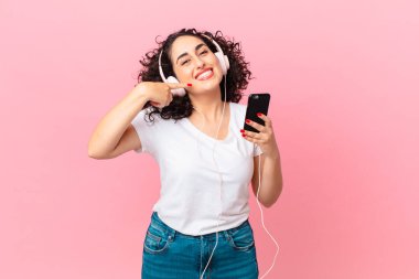 Güzel Arap kadın kendine güvenerek gülümsüyor kulaklıkları ve akıllı telefonuyla kendi gülüşünü işaret ediyor.