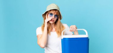 Güzel, kızıl saçlı bir kadın yorgun ve uykulu ve sıkılmış hissediyor ve elinde taşınabilir bir buzdolabı tutuyor. yaz konsepti