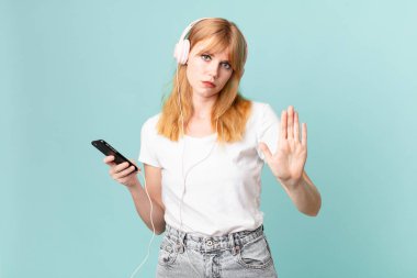 Güzel kızıl saçlı kadın ciddi görünüyor. Avuç içini gösteriyor. Durma hareketi yapıyor ve kulaklıkla müzik dinliyor.