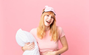Güzel kızıl saçlı kadın komik bir şakaya yüksek sesle gülüyor. Pijama giyiyor ve yastık tutuyor.