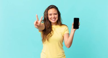 Genç ve güzel bir kadın gülümsüyor ve mutlu görünüyor. Zafer ya da barış işareti yapıyor ve elinde akıllı bir telefon tutuyor.