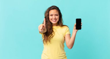 Genç ve güzel bir kadın gururla gülümsüyor ve kendine güvenerek bir numara oluyor ve elinde akıllı bir telefon tutuyor.