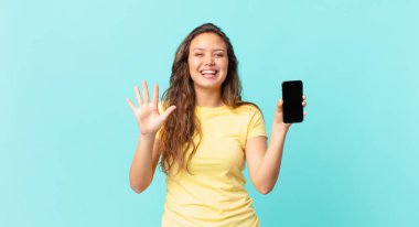 Genç, güzel bir bayan gülümsüyor ve arkadaş canlısı görünüyor. Beş numarayı gösteriyor ve elinde akıllı bir telefon var.