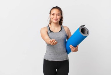 Genç ve güzel bir kadın mutlu bir şekilde gülümsüyor ve bir konsept sunuyor ve yoga minderi tutuyor.