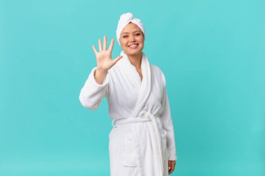 Genç, güzel bir kadın gülümsüyor ve arkadaş canlısı görünüyor. Beş numarayı gösteriyor ve duştan sonra banyo cübbesi giyiyor.