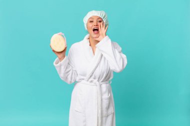 Genç, güzel bir kadın kendini mutlu hissediyor. Elleri ağzının yanında ve duştan sonra banyo cübbesi giyiyor.