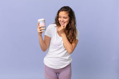 Genç, güzel bir kadın, mutlu, kendinden emin bir ifadeyle gülümsüyor. Elleri çenesinde ve elinde kahve tutuyor.