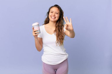 Genç, güzel bir bayan gülümsüyor ve arkadaş canlısı görünüyor. Üç numarayı gösteriyor ve elinde kahve tutuyor.