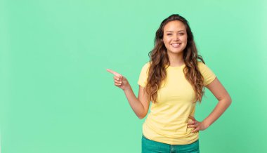 Genç ve güzel bir kadın mutlu bir şekilde gülümsüyor, eli kalçasında, kendinden emin ve fotokopi alanını işaret ediyor.