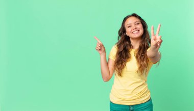 Genç ve güzel bir kadın gülümsüyor ve mutlu görünüyor. Zafer ya da barış işareti yapıyor ve fotokopi alanını işaret ediyor.